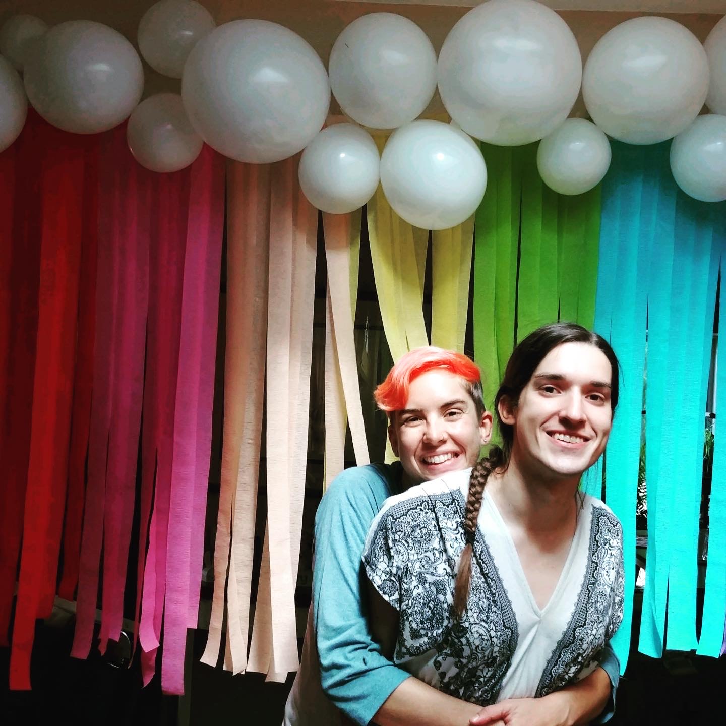 Mel and Teghan hugging in front of rainbow streamers